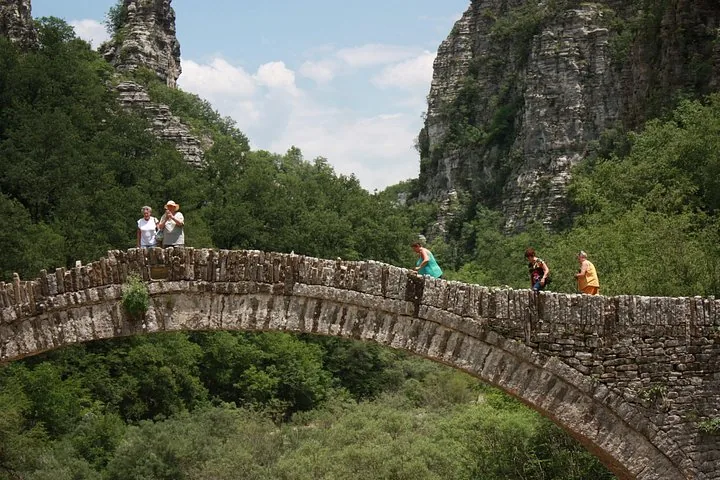 People walking across an old stone arch bridge in nature.